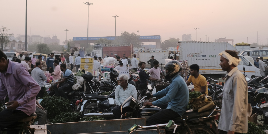 Busy street scene in Delhi with heavy car traffic, pedestrians, and street vendors navigating a crowded, bustling urban environment.